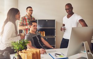 Diverse group of multi-ethnic young adult business people assembled around desk looking at something on their computer monitor