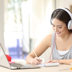 student wearing headphones while writing and working in front of a laptop