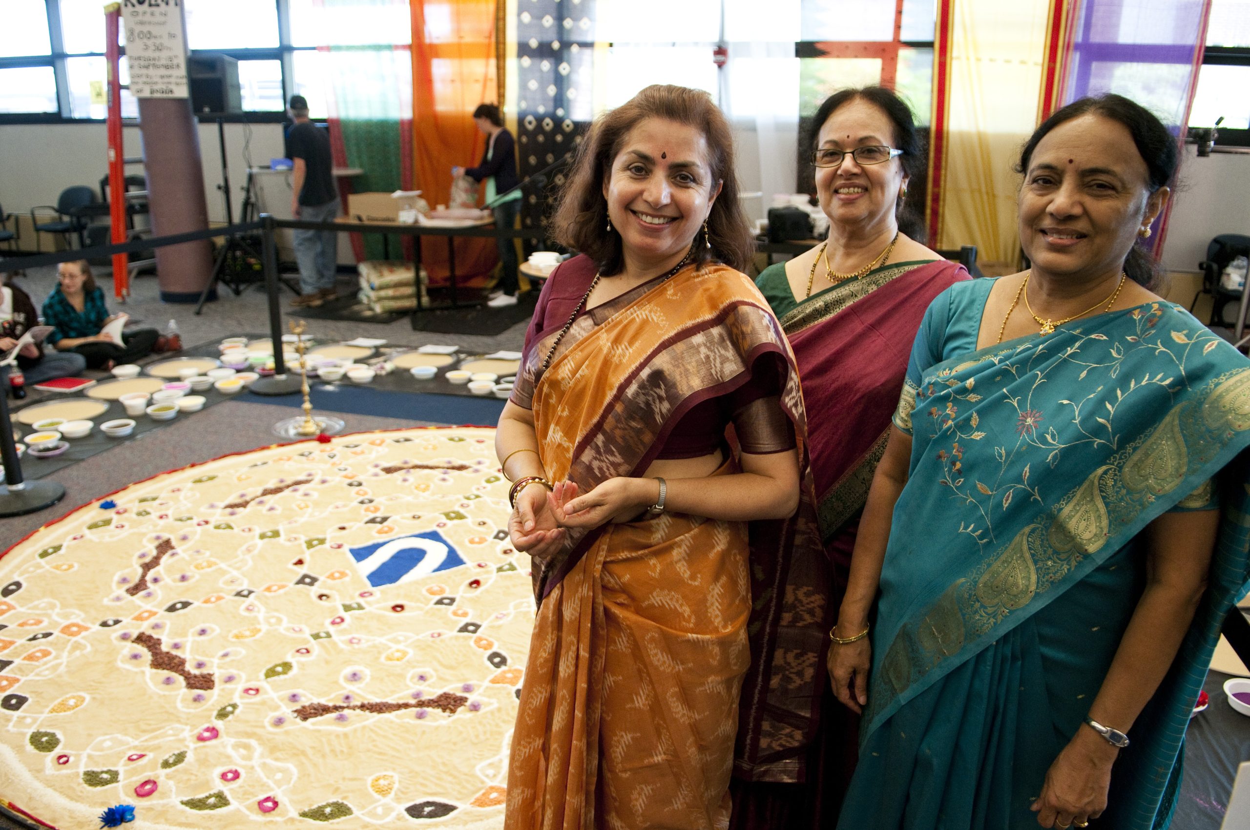 Women dressed in traditional Indian Saris at Northampton Community College pose for a picture in front of a Kolam art image of NCC’s logo